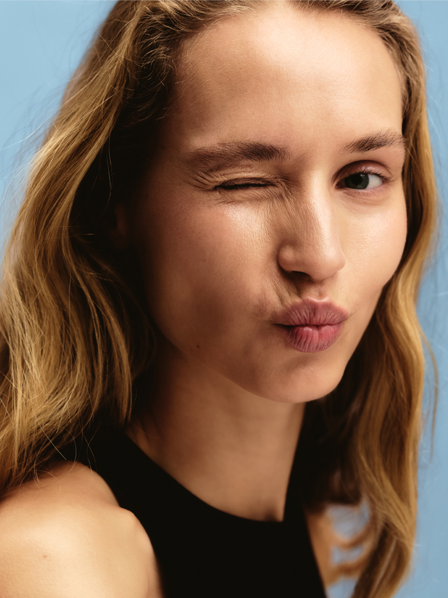 Young woman with long wavy red hair making a playful pout and winking, wearing a black top against a light blue background.