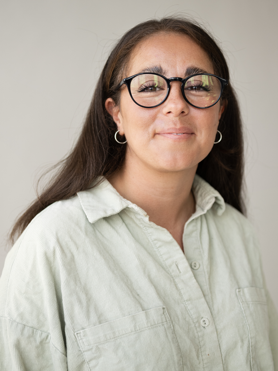 A woman with long brown hair, wearing black glasses, hoop earrings, and a light-colored button-up shirt, standing against a plain background.