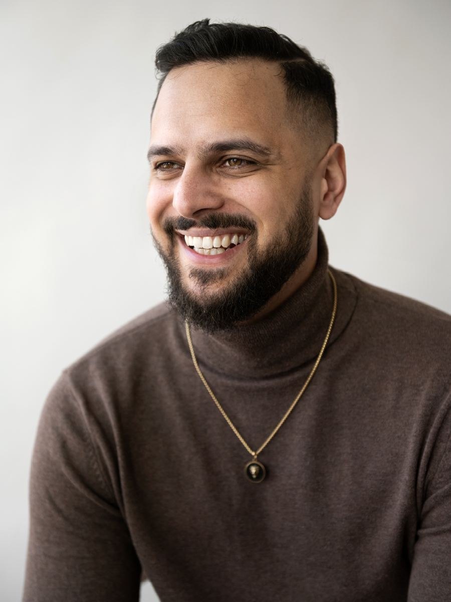 A smiling man with a beard and short dark hair, wearing a brown turtleneck sweater and a gold necklace with a circular pendant, against a plain light background.
