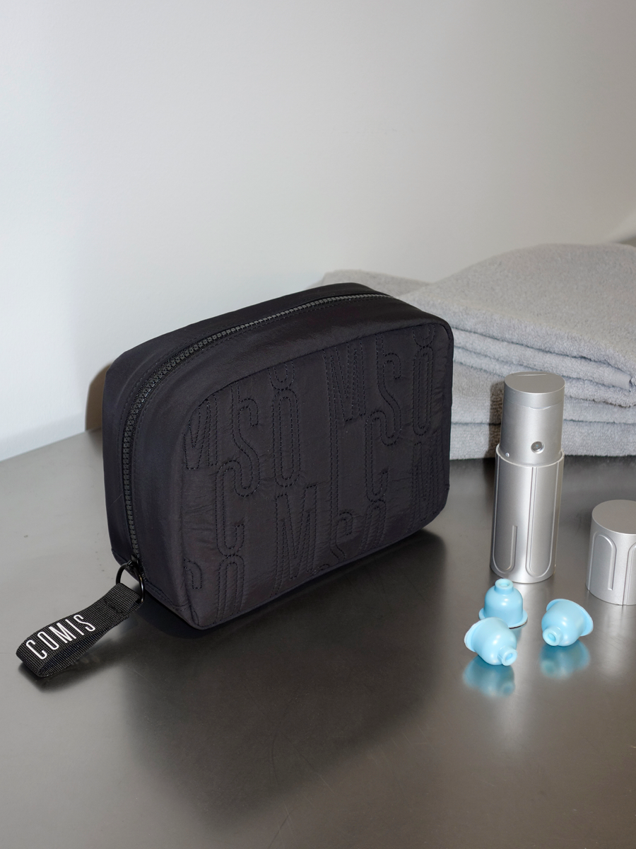 A black zippered toiletry bag with a black handle tagged 'COMMS' is on a gray surface next to a set of light gray towels, a silver spray bottle, a small silver container, and three blue earbuds.