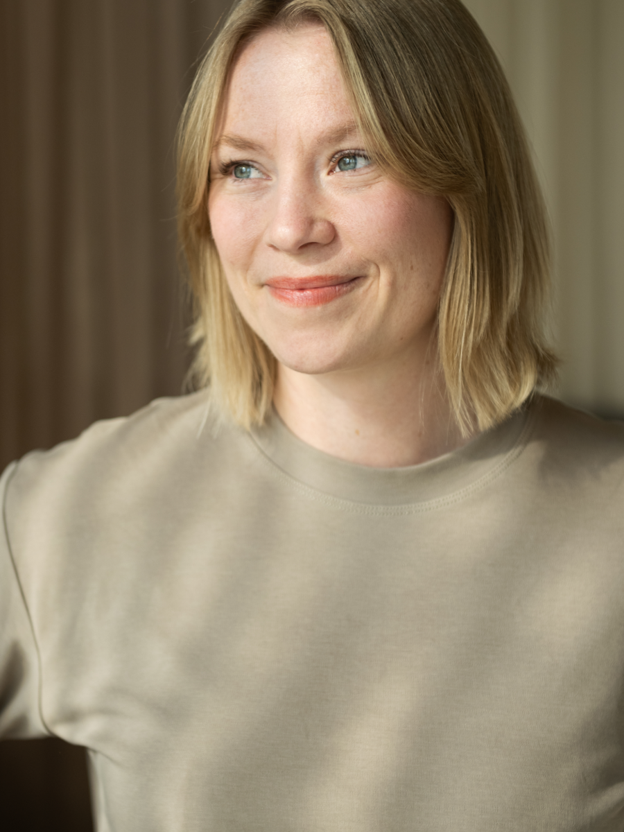 A smiling woman with shoulder-length blonde hair, wearing a light beige shirt, standing indoors near a neutral-colored background.