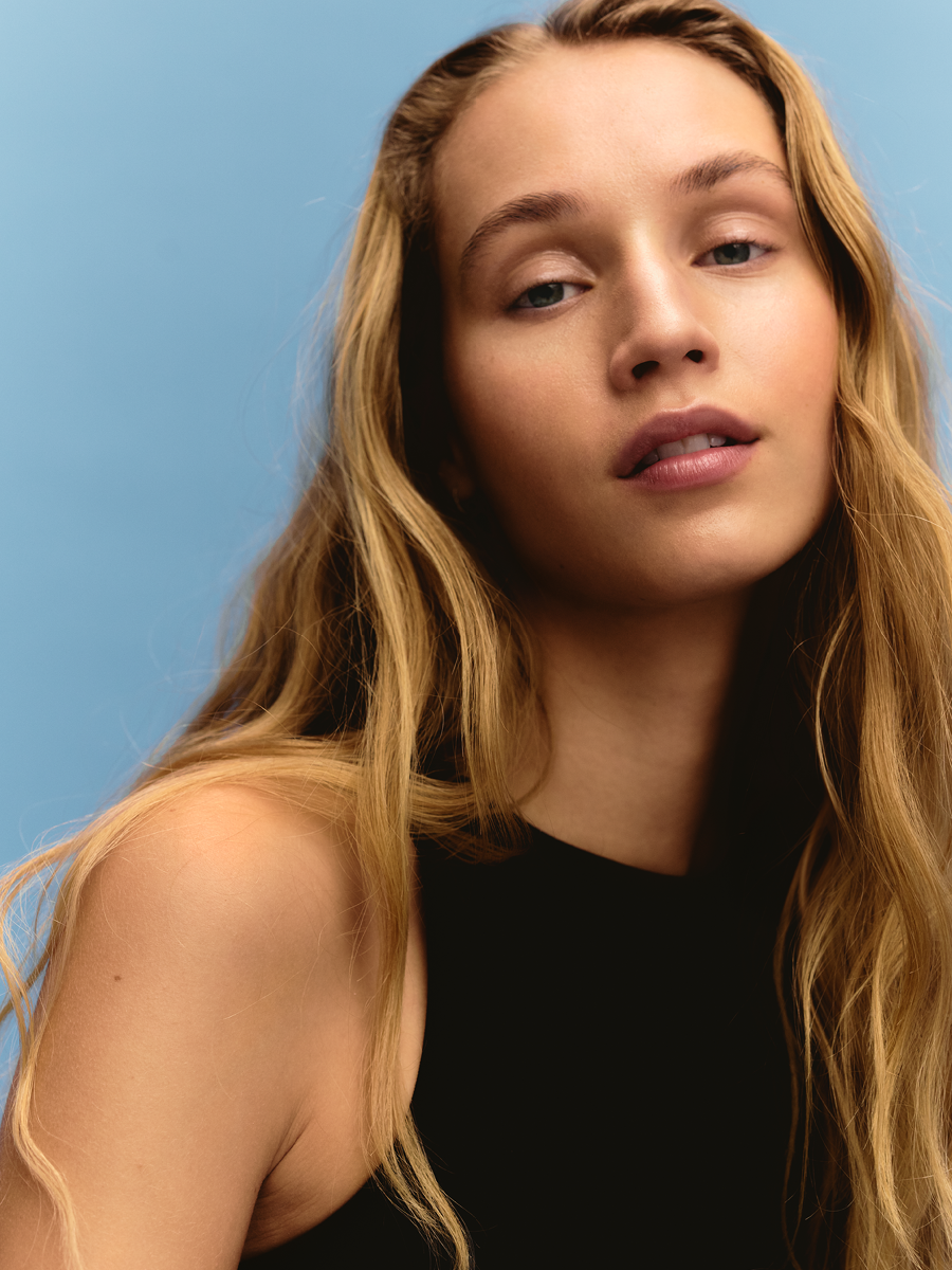 Close-up portrait of a young woman with long wavy blonde hair, wearing a black sleeveless top, against a blue background.