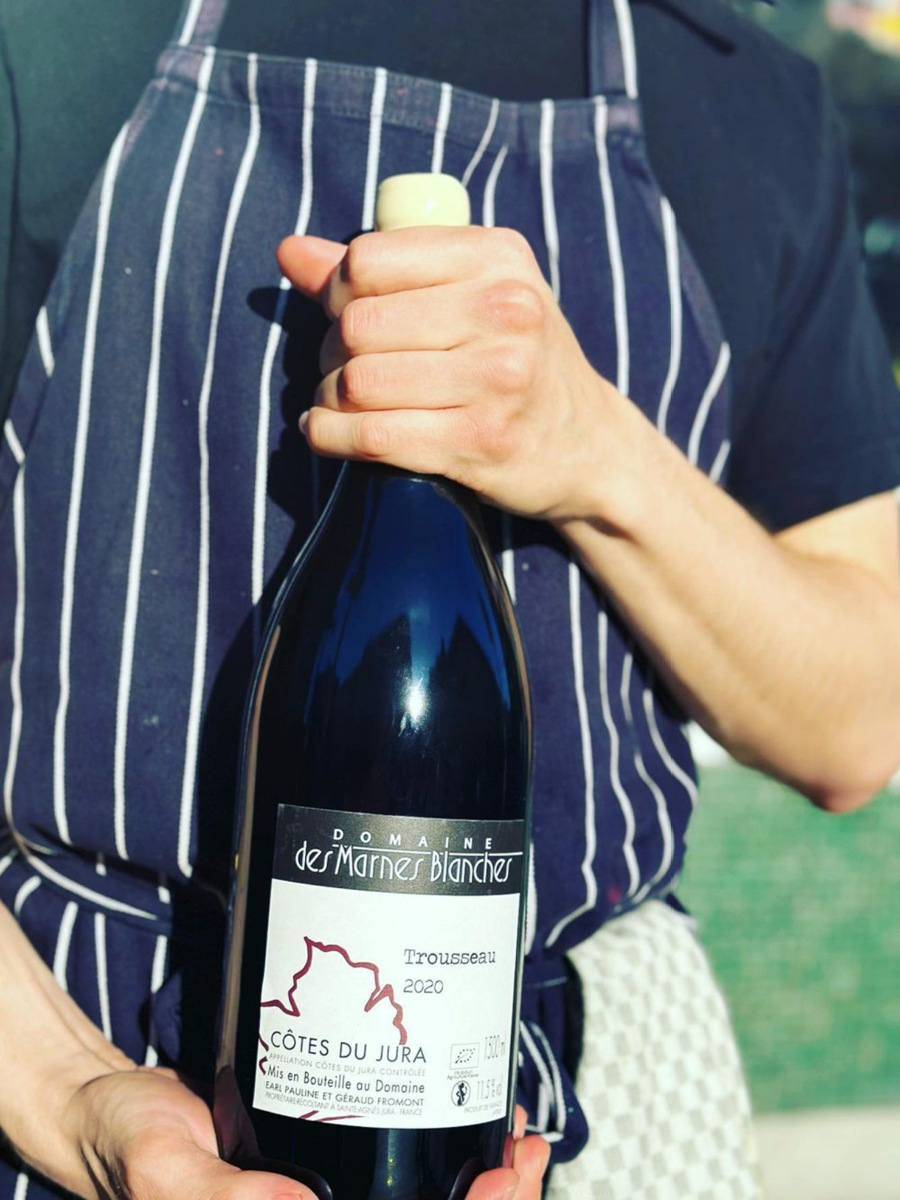 Person wearing a dark blue and white pinstripe apron holding a bottle of Domaine des Marines Blanches Trousseau 2020 wine from Côtes du Jura, in an outdoor setting.