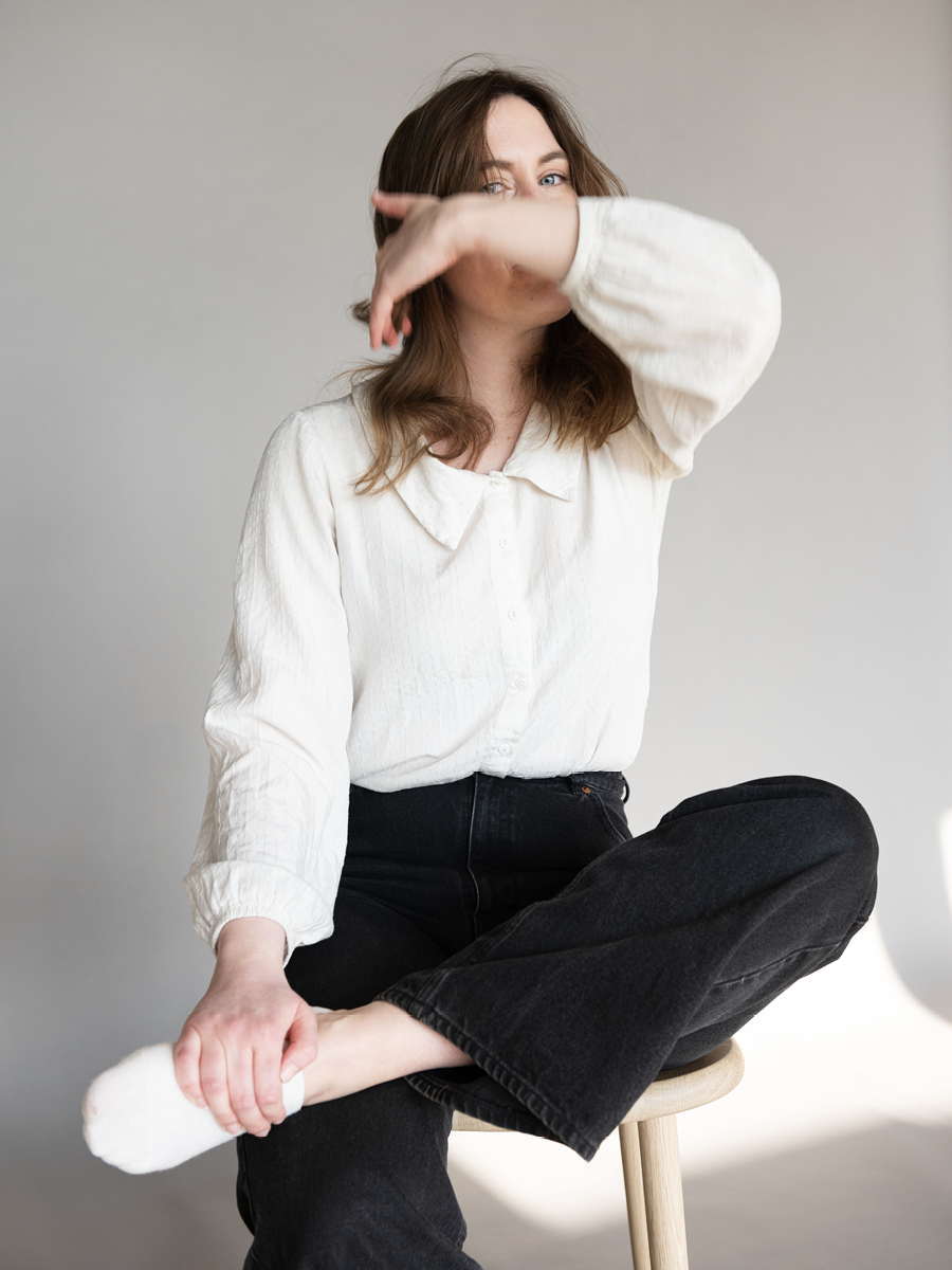 A woman with brown hair and blue eyes sitting cross-legged on a chair against a plain background, covering part of her face with her left arm.