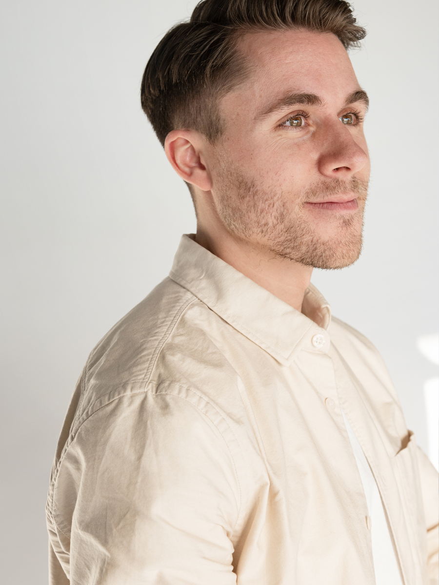 A young man with brown hair and light skin, wearing a beige button-up shirt, looking slightly to the right with a calm expression.