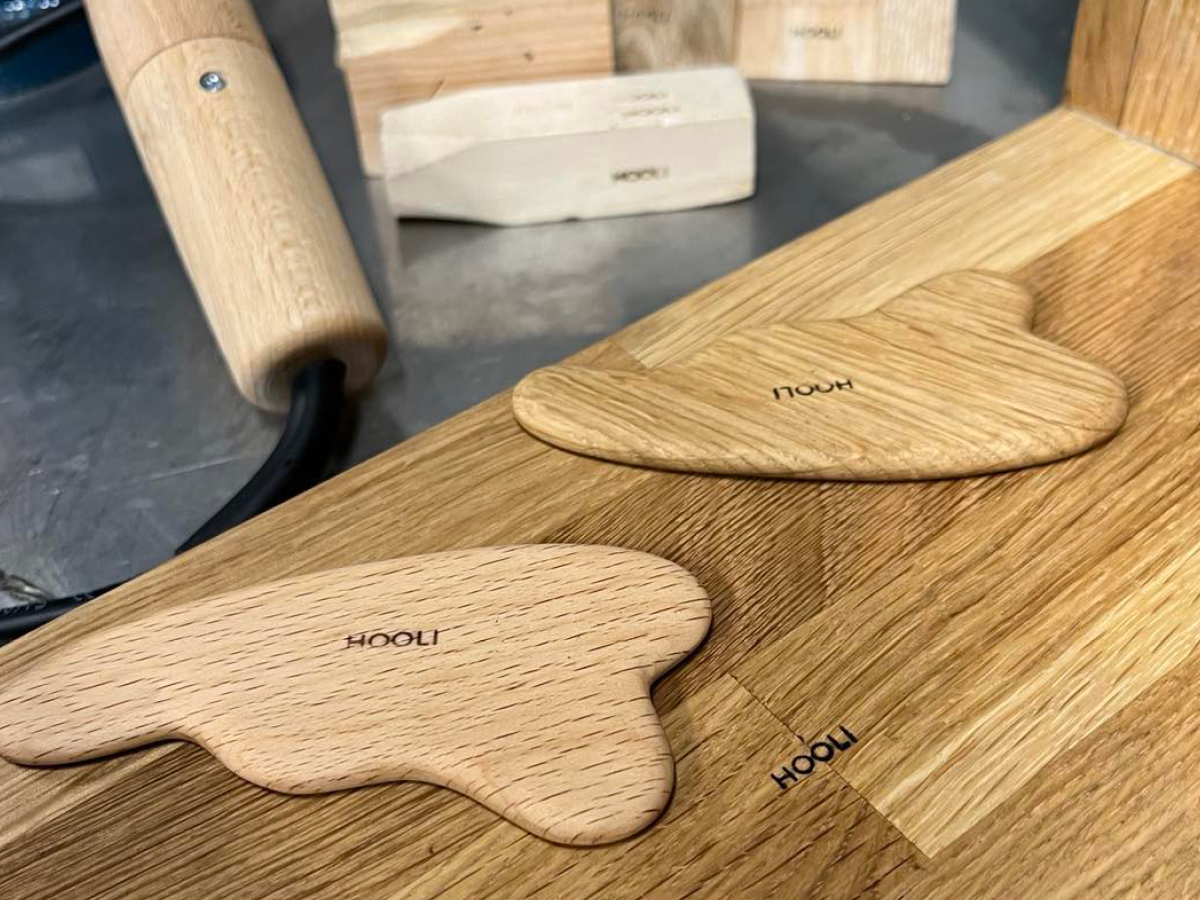 Close-up of two wooden paddle-shaped pieces with the word 'HOOH' stamped on each, resting on a wooden surface, with a blurred background of woodworking tools.