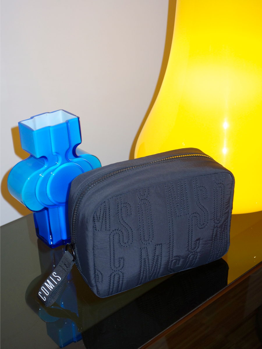 A blue plastic vase, a black zippered pouch with the word 'COS' embroidered on it, and a yellow lamp on a glass surface.