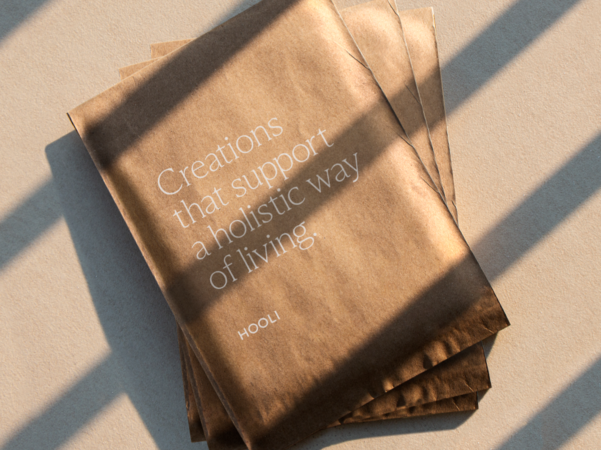Stack of brown paper bags with white text reading, 'Creations that support a holistic way of living,' placed on a beige surface with shadows from a window.