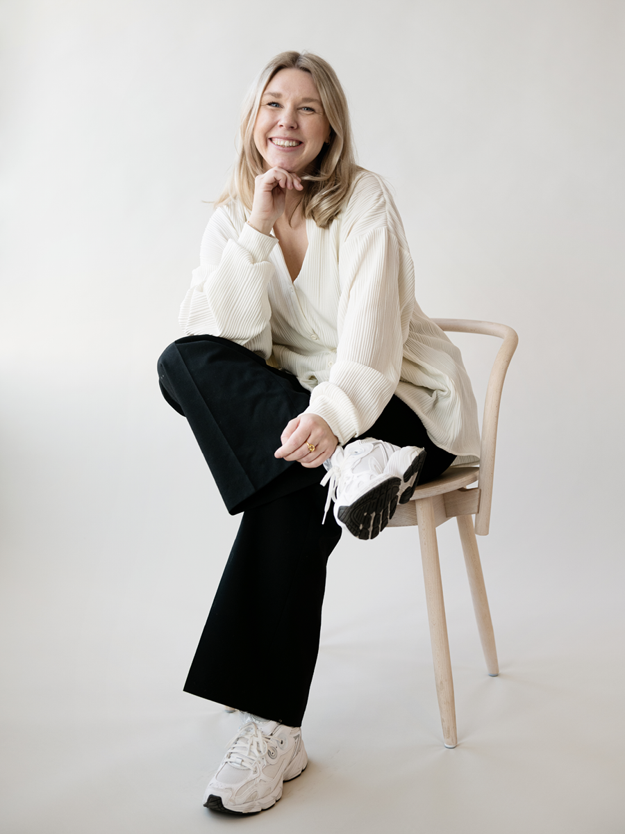A woman with blonde hair, smiling, sitting on a wooden chair with one leg crossed over the other, wearing white sneakers, black pants, and a cream-colored blouse against a plain white background.