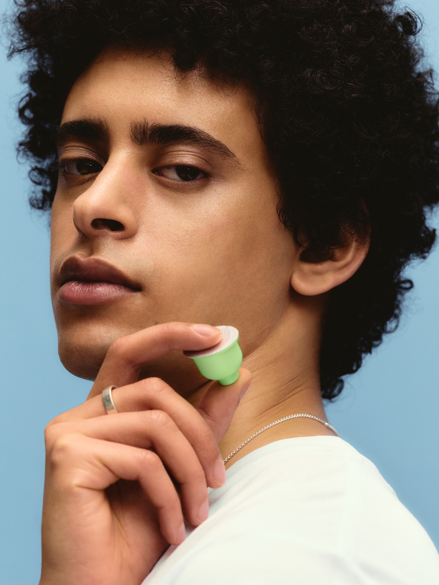 A young person with curly black hair holding a green facial roller near their face against a light blue background.