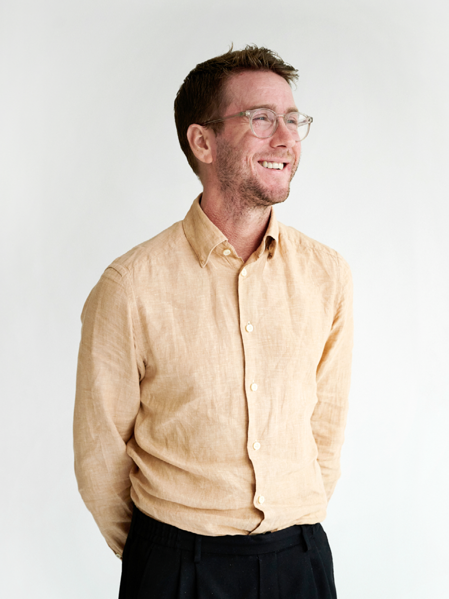 Smiling man with glasses and a beard, wearing a beige button-up shirt and black pants, standing against a plain white background.