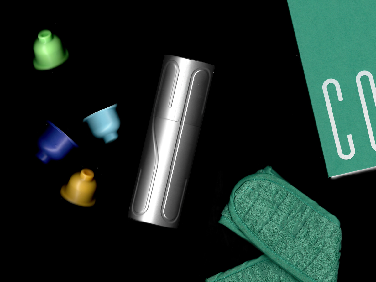A silver electronic vaping device, four colorful cylindrical vape cartridges in green, blue, yellow, and light blue, a teal towel with the words "VAPE SHOP" embossed, and a green cardboard box partially visible.