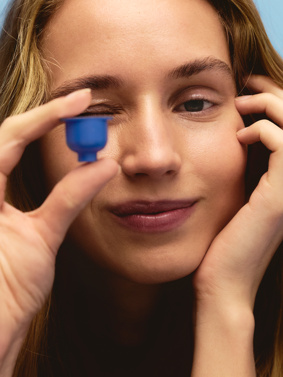 A woman with light skin and brown hair holds a blue contact lens case near her eye, preparing to put in a contact lens.