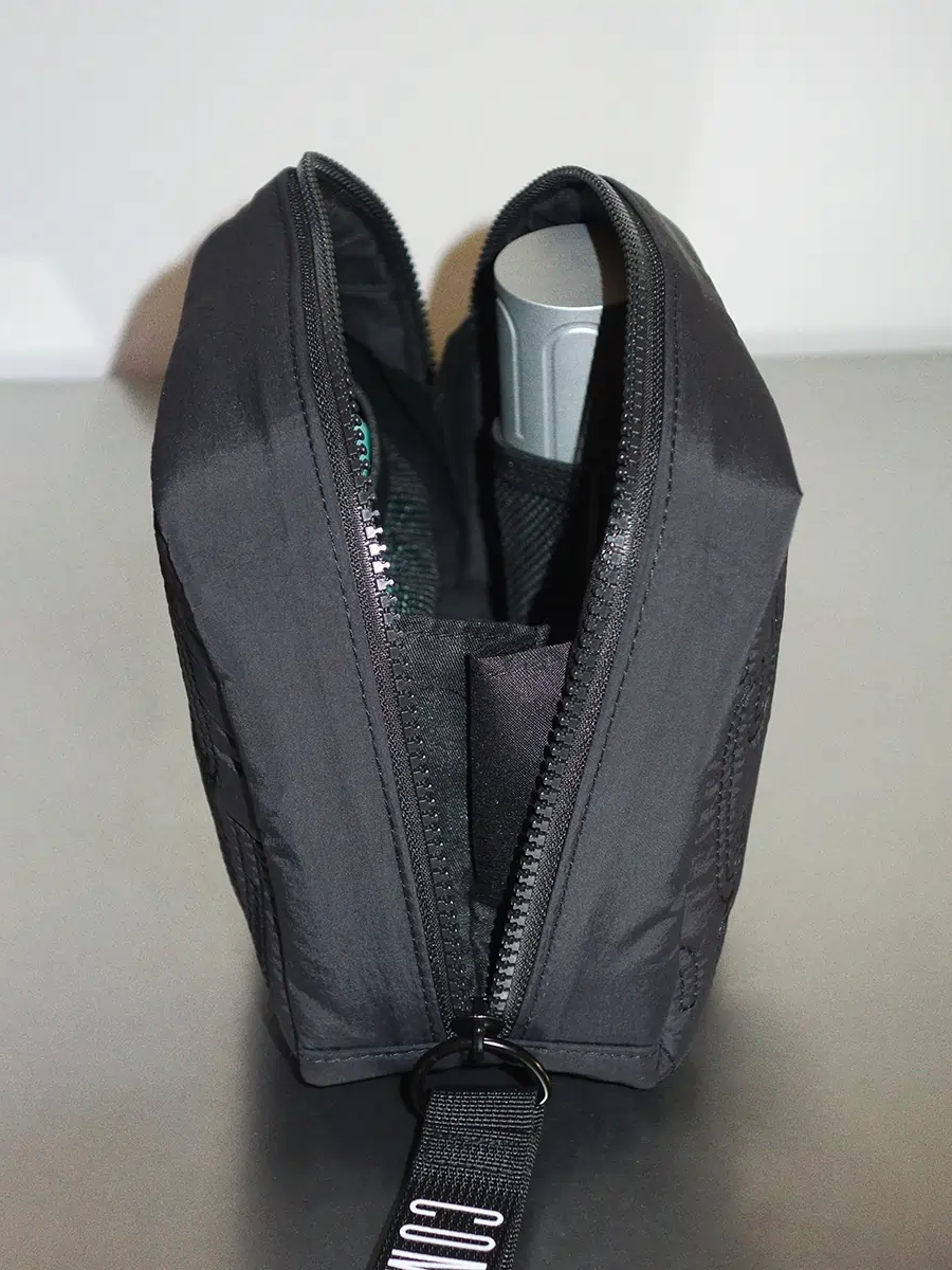 Black zippered pouch with items inside, including a gray spray can, placed on a gray surface