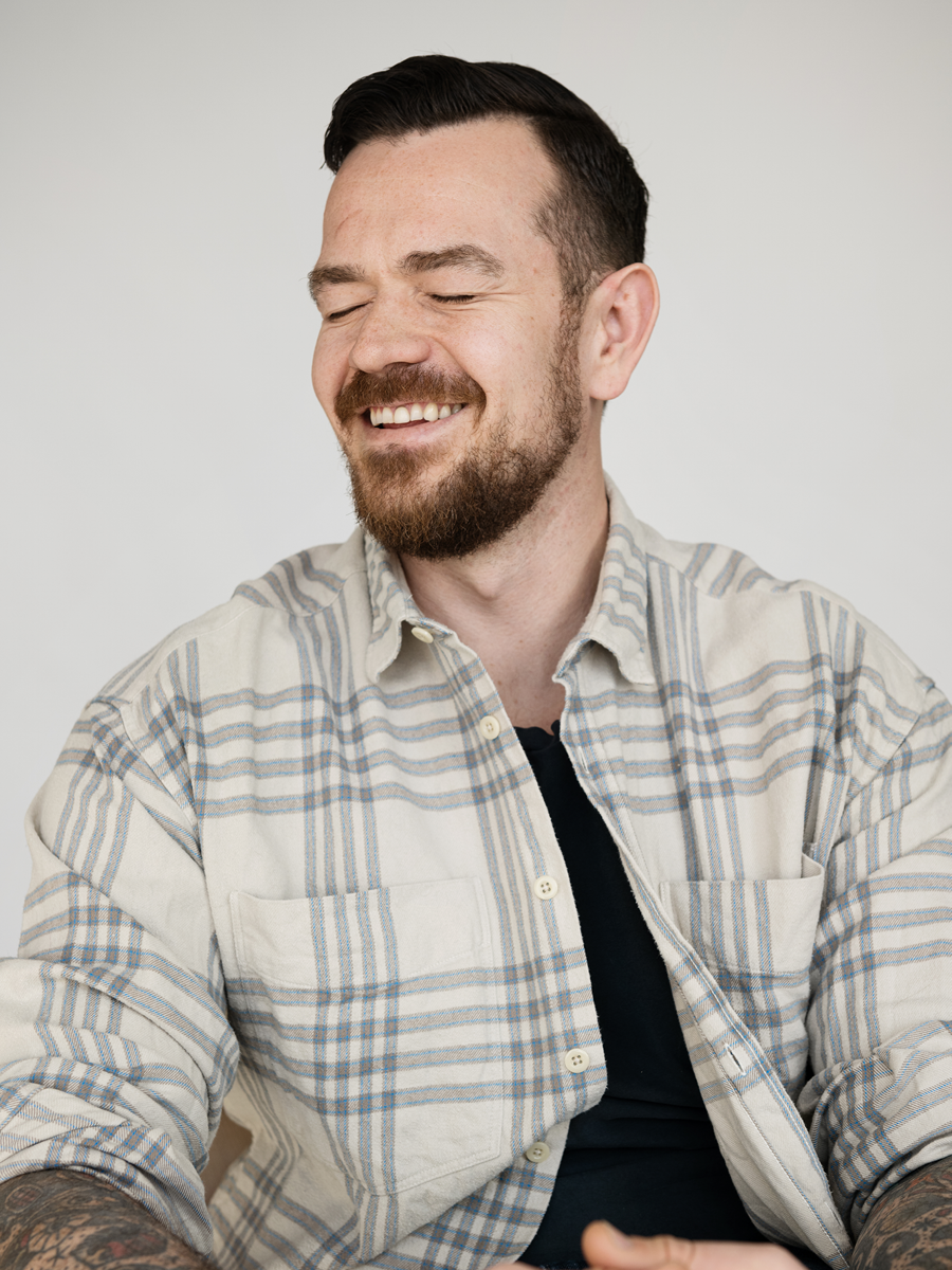 A smiling man with a beard and tattoos, wearing a plaid shirt and black t-shirt, with his eyes closed and head slightly turned to the side.