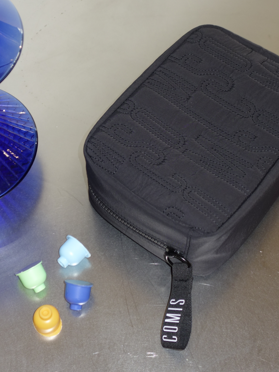 A black zipper case labeled 'SIX COM' with a black handle attached, placed on a gray surface next to four colorful headphone ear tips in green, blue, yellow, and white, and partially visible blue glass objects.