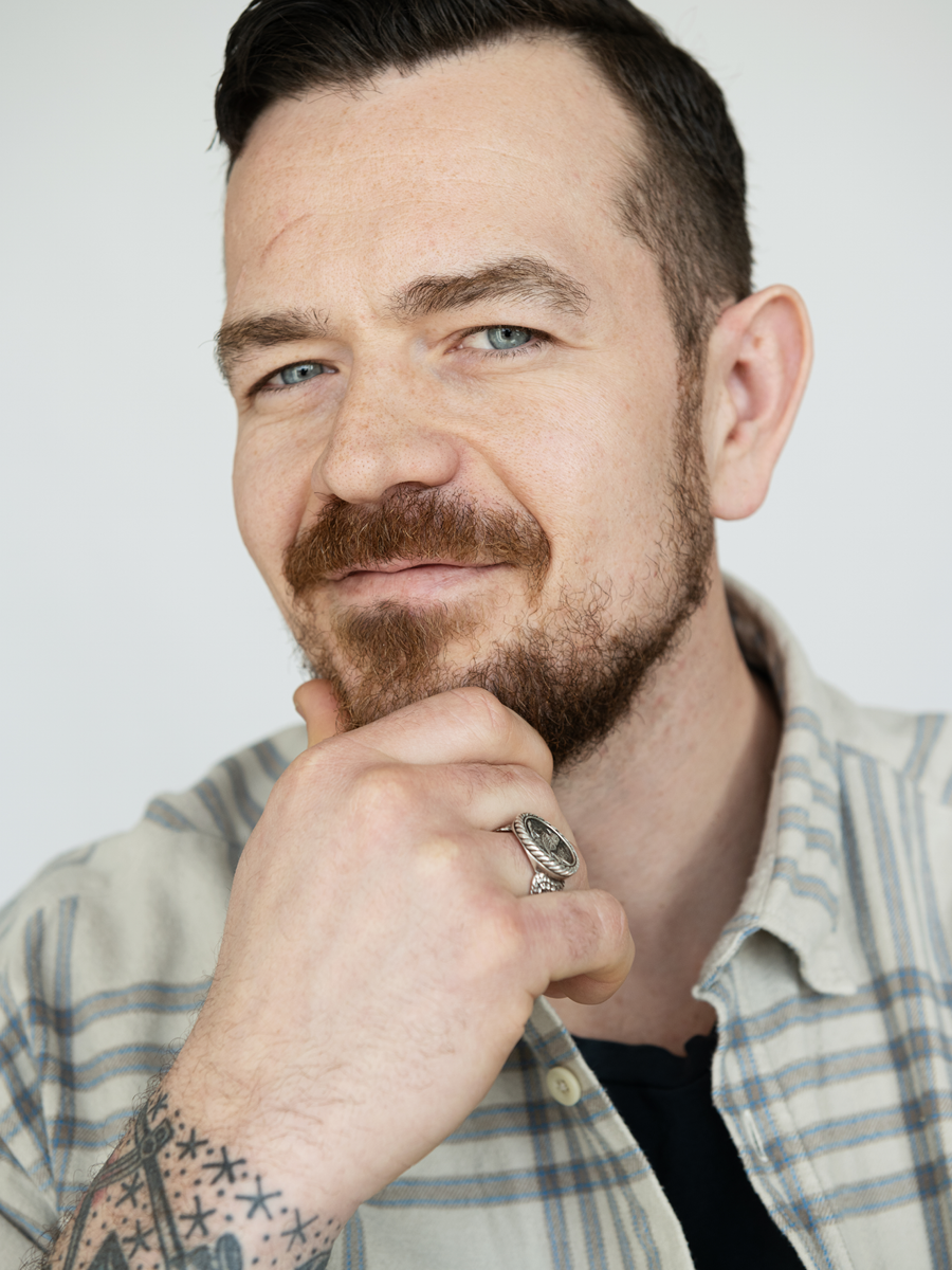 Close-up of a man with a beard and mustache, wearing a plaid shirt, resting his chin on his hand, with a silver ring on his finger, and visible tattoos on his wrist.