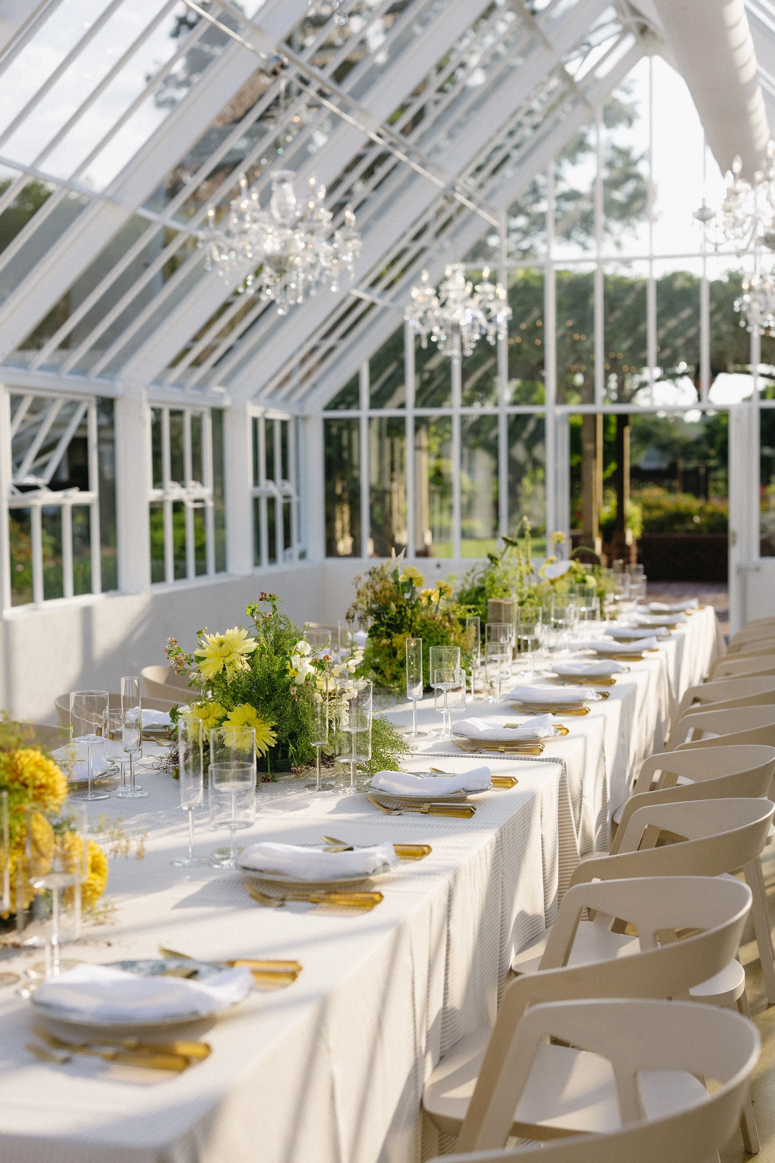 Elegant outdoor wedding reception table decorated with white flowers, tall candles in glass holders, and clear chairs in a garden setting.