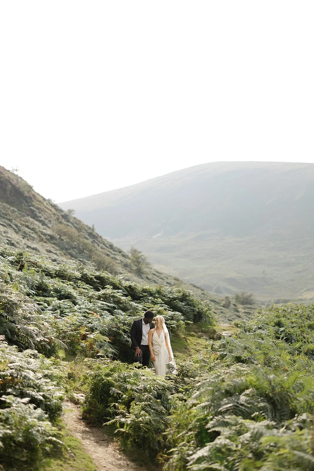 A couple walking hand in hand through a lush, mountainous landscape with green foliage and distant hills.
