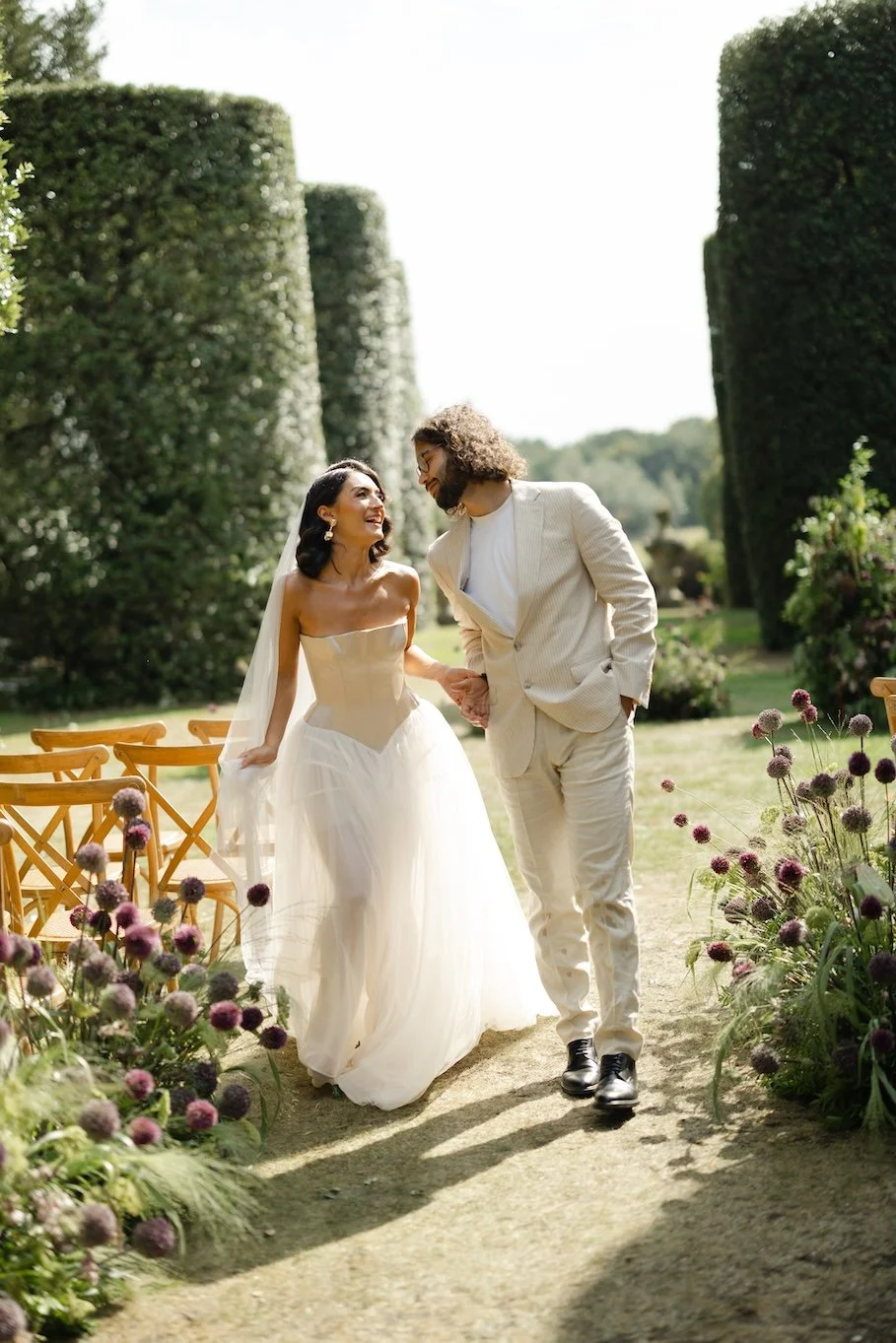 A newlywed couple holding hands and walking outdoors on a sunny day, surrounded by purple flowers and green hedges, smiling at each other.