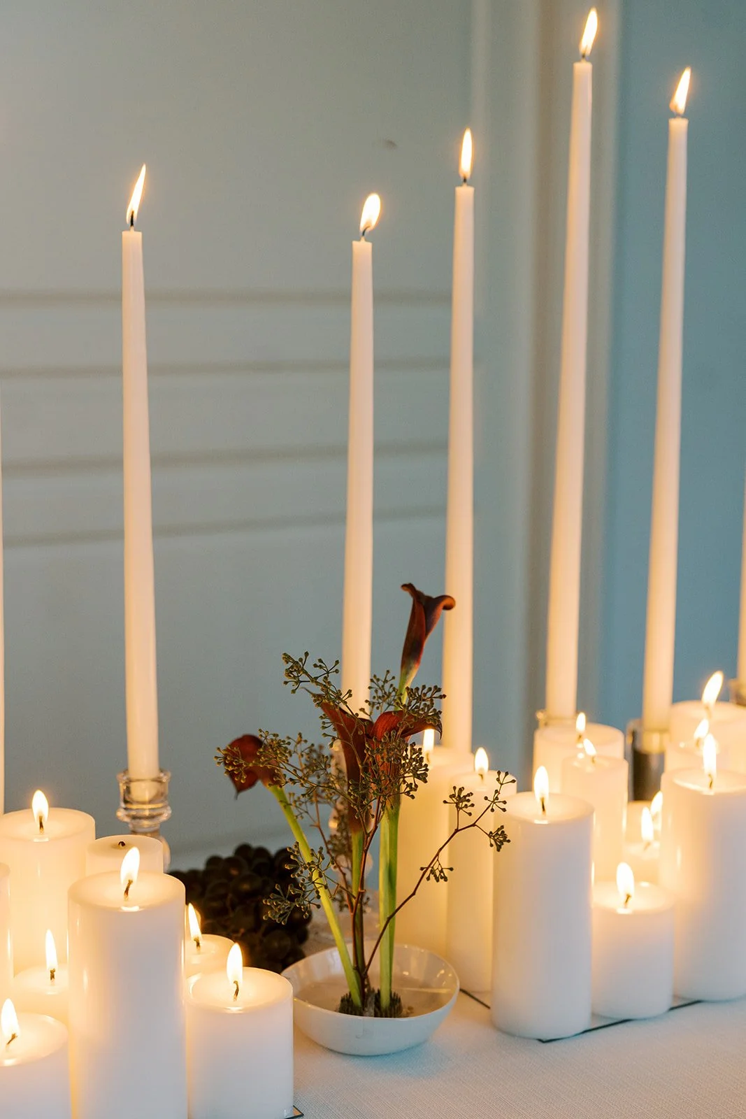 A centerpiece with dark red calla lilies and greenery surrounded by white candles of various heights, all lit, on a white tablecloth.