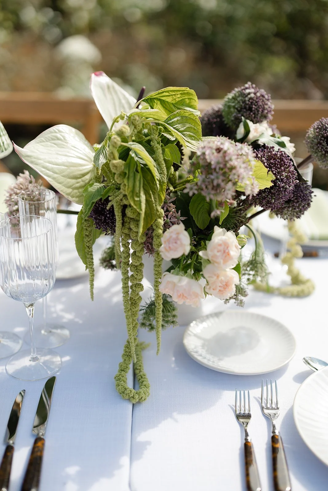 Elegant table setting with a floral centerpiece featuring pink and purple flowers with green leaves, set on a white tablecloth with glassware, plates, and silverware.