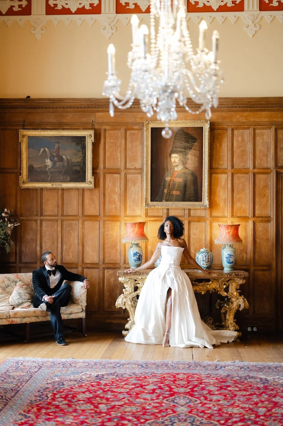 A woman in an elegant white dress posing in a luxurious room with wood-paneled walls. A man in a tuxedo sits on a floral sofa, watching her. The room features portraits, antique vases, and ornate furniture, with a chandelier hanging from the ceiling.