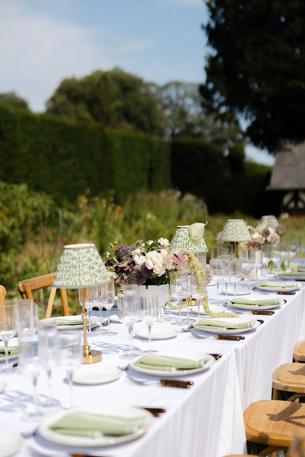 Elegant outdoor dining table set with white tablecloth, floral centerpieces, green lamps, glassware, plates, and cutlery on a sunny day in a garden.