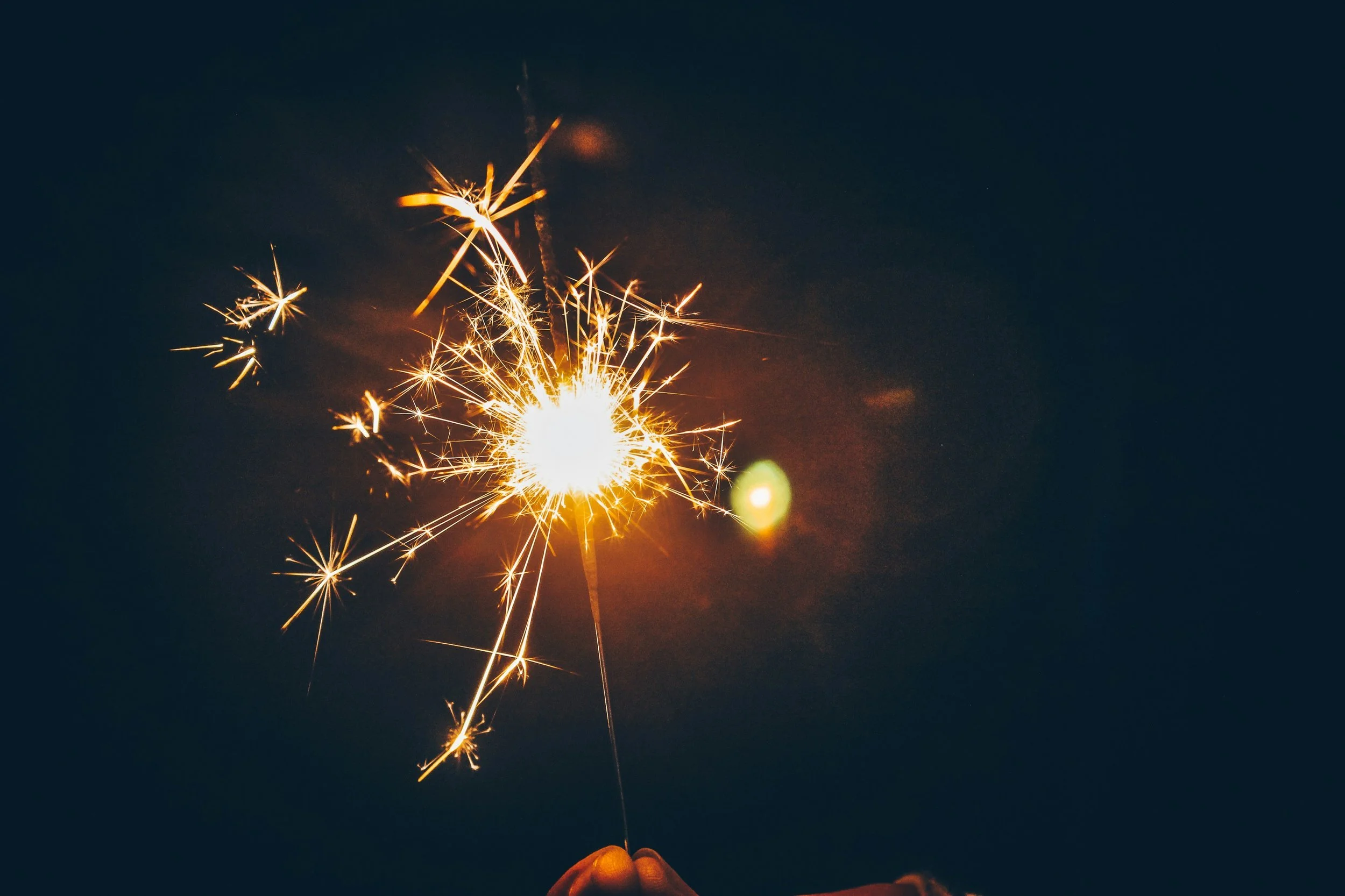 Person holding a lit sparkler with bright sparks in a dark background.