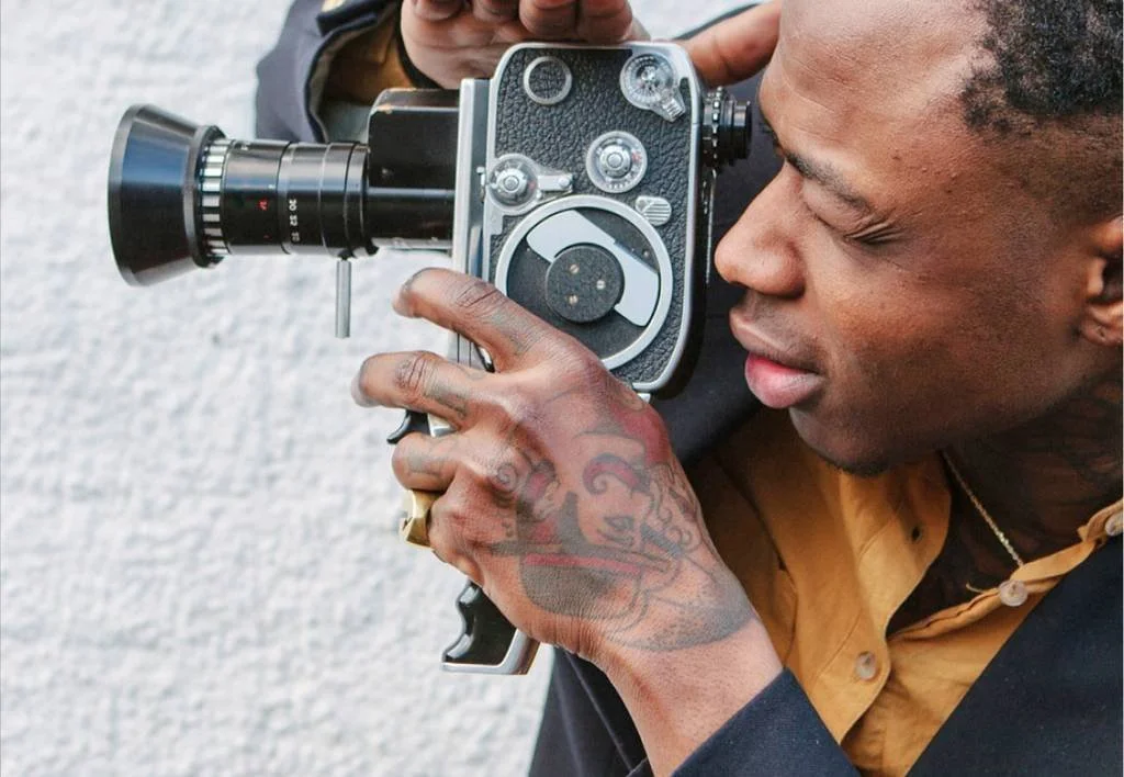 Man with tattoos on hand using a vintage camera with a large lens, focusing to take a picture.