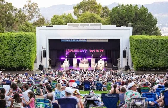 Outdoor concert with a large audience seated on lawn chairs in front of a stage surrounded by trees and mountains.
