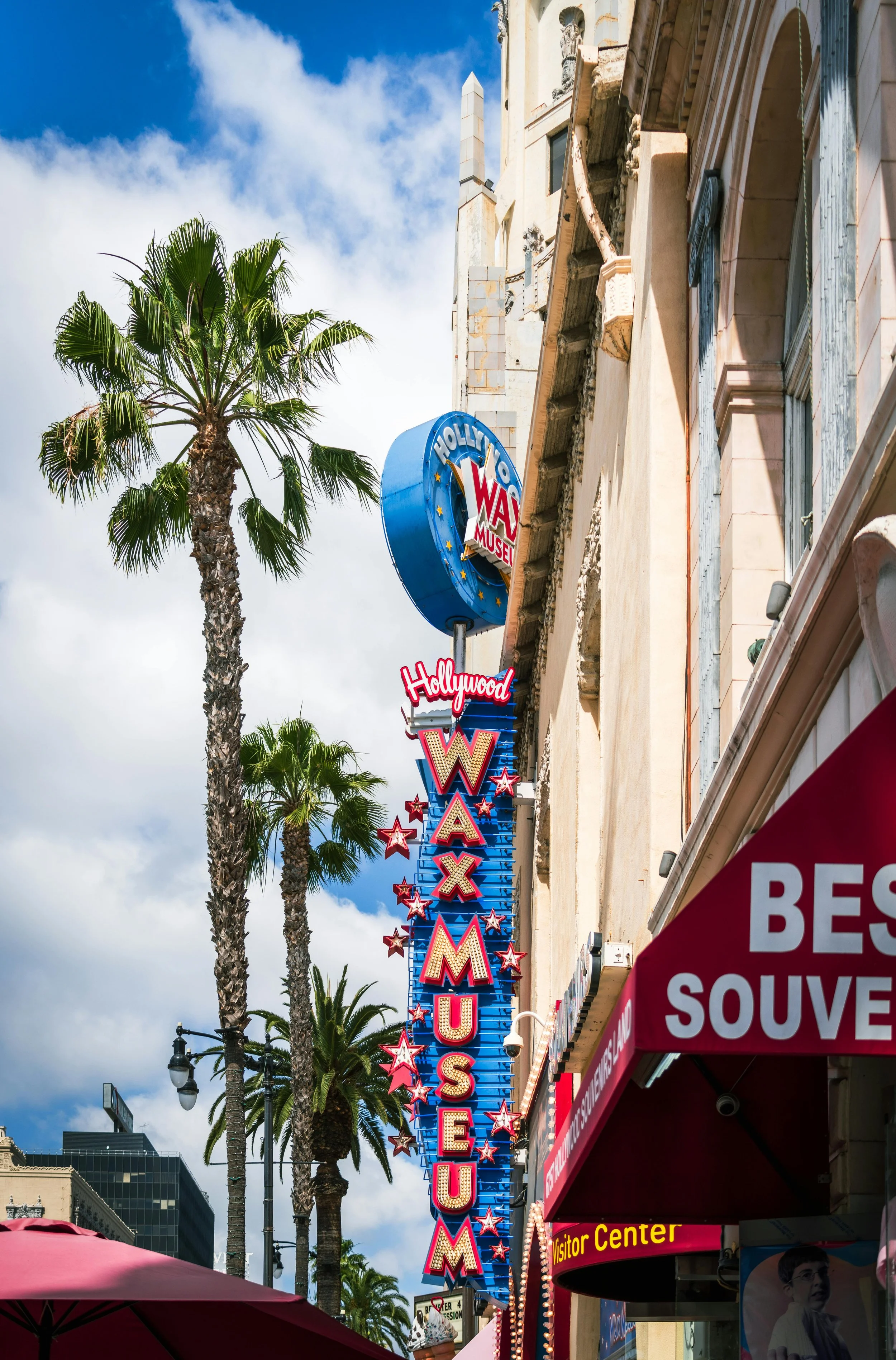 The iconic Hollywood WAX Museum sign with palm trees, a partly cloudy sky, and a red awning in the foreground.