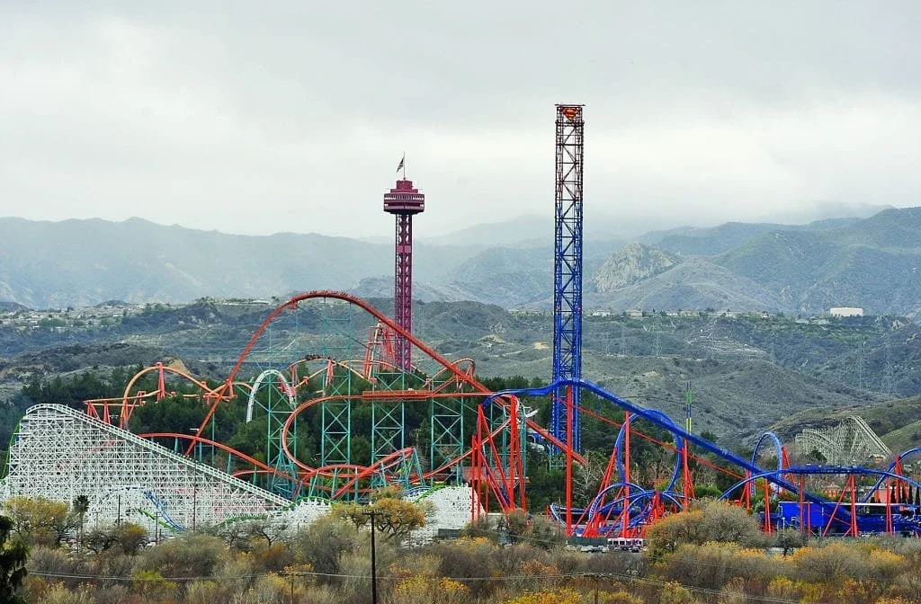 Colorful amusement park with roller coasters and a tower, set against mountainous backdrop under overcast sky.