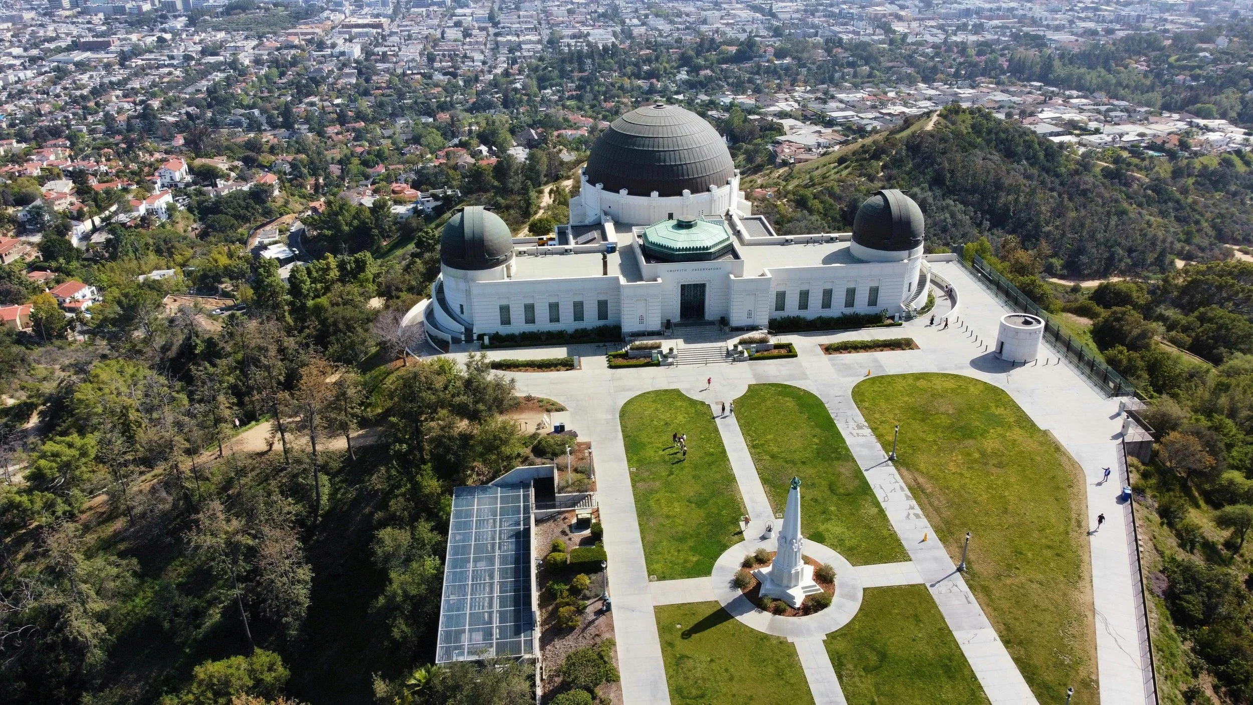 Aerial view of the Griffith Observatory situated on a hill, overlooking Los Angeles residential area and cityscape, with green lawns, pathways, and a monument in front.