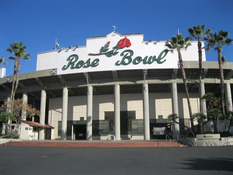 Exterior view of the Rose Bowl stadium with tall palm trees, large columns, and the sign 'Rose Bowl' with a rose graphic at the top.
