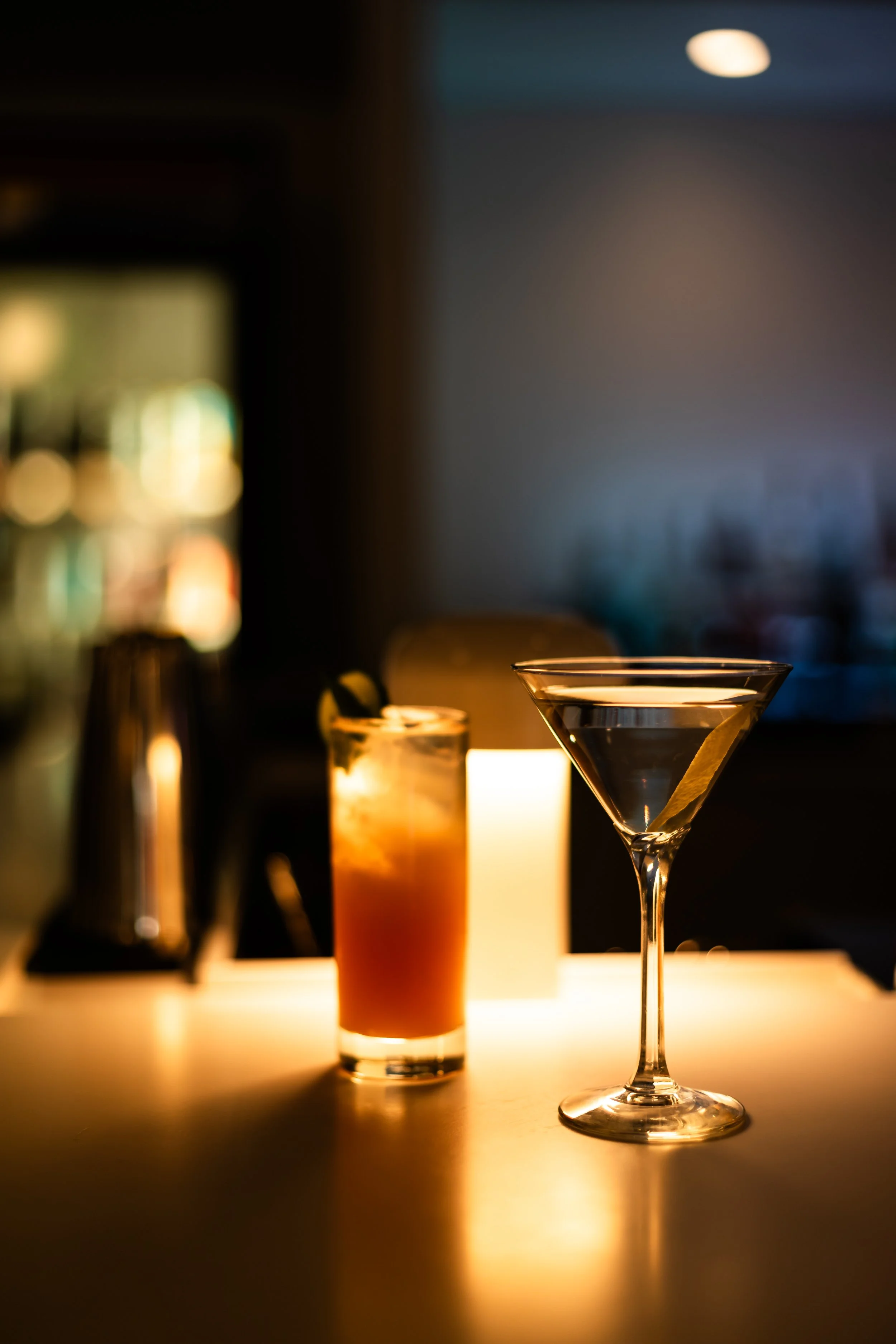 Three cocktails on a bar table illuminated by a candle, with a blurred background.