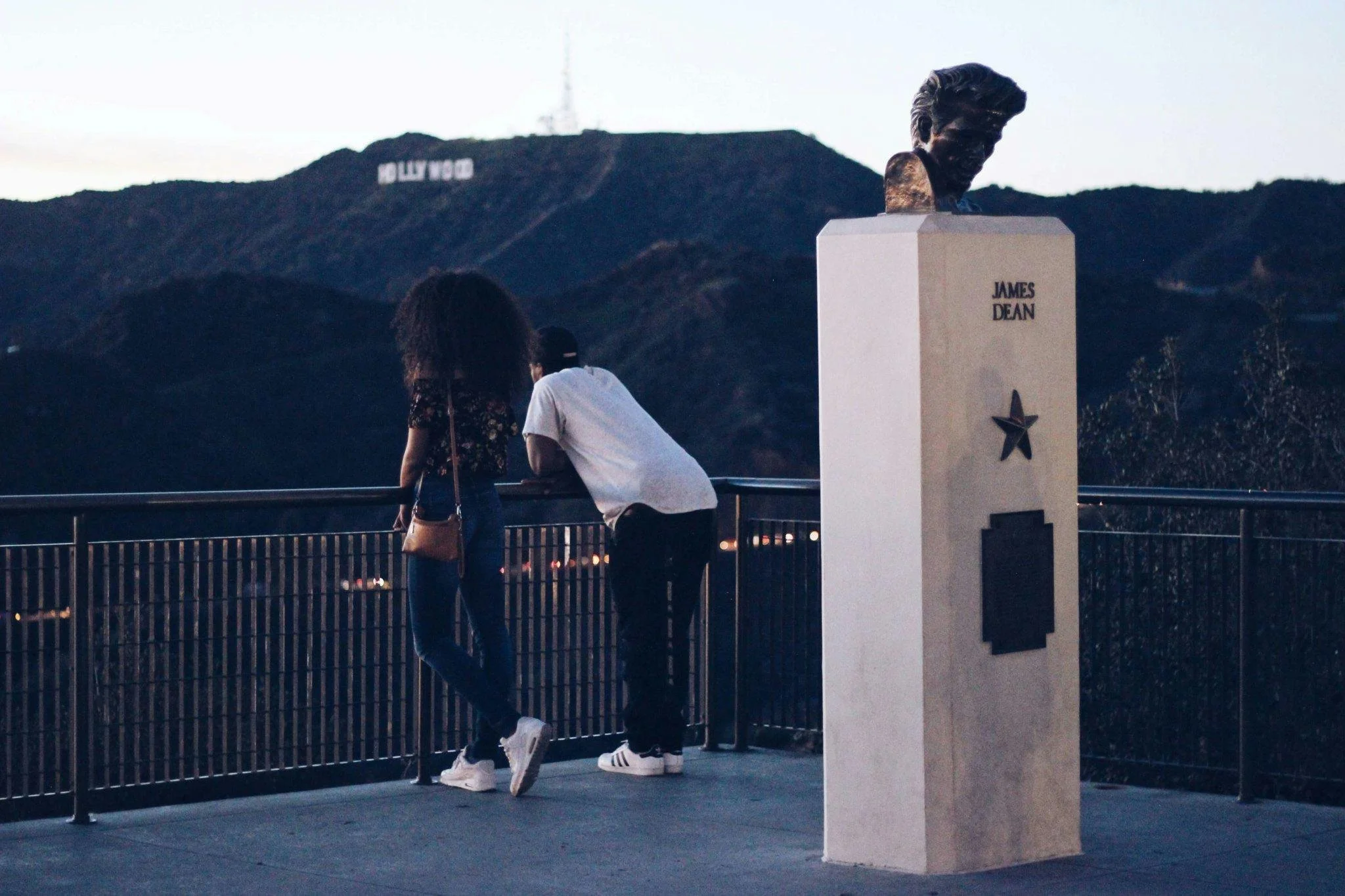 Two people looking out from the Griffith Observatory in Los Angeles, with the Hollywood Sign on the hillside in the background at dusk.