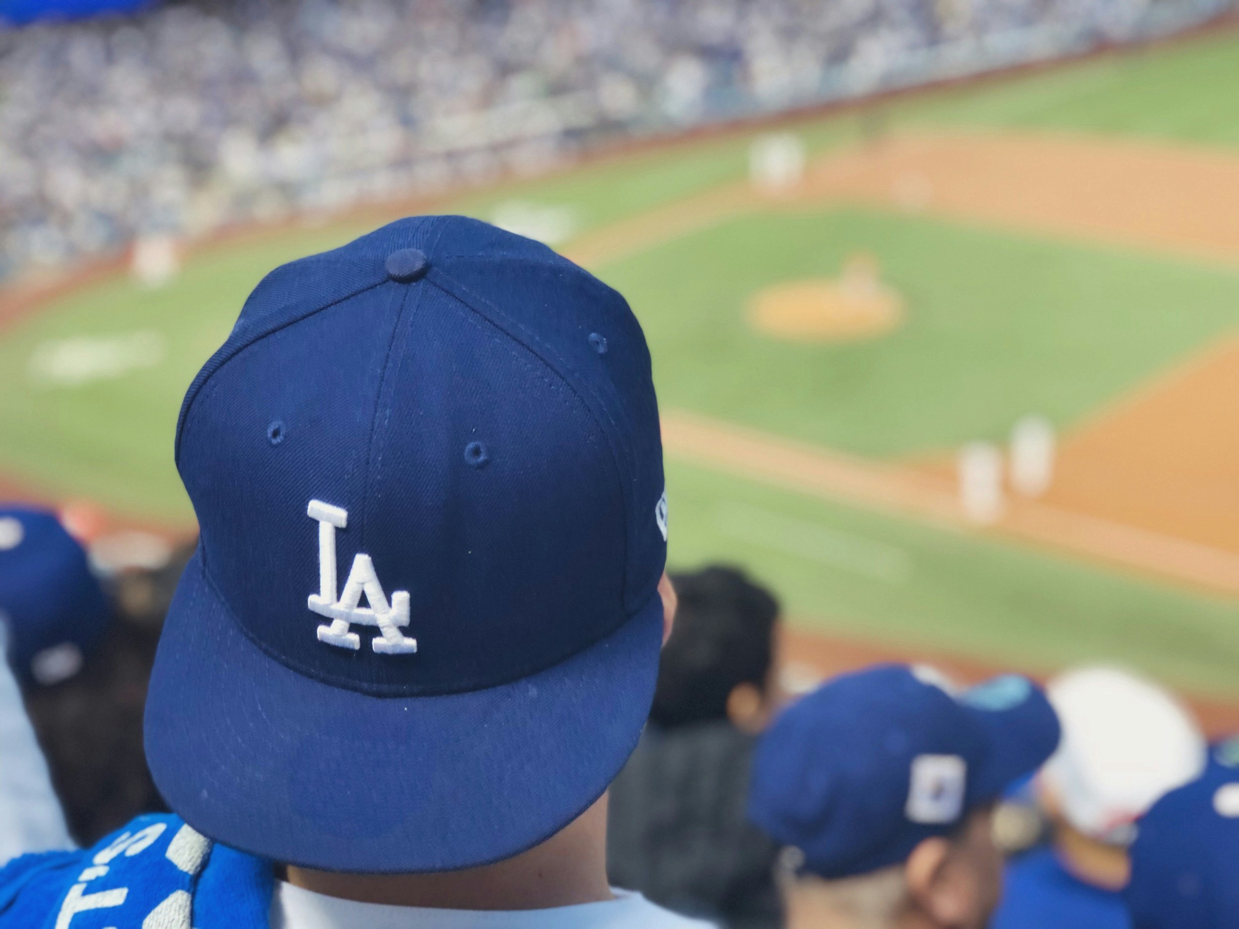 Close-up of a person wearing a blue Los Angeles Dodgers baseball cap at a baseball stadium, with a view of the field and other spectators in the background.