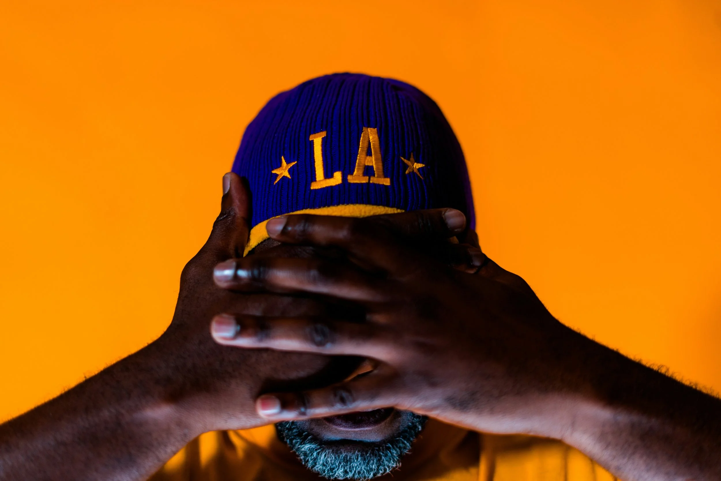 A person with dark skin and a gray beard covering their eyes with both hands, wearing a blue and yellow LA beanie, against an orange background.