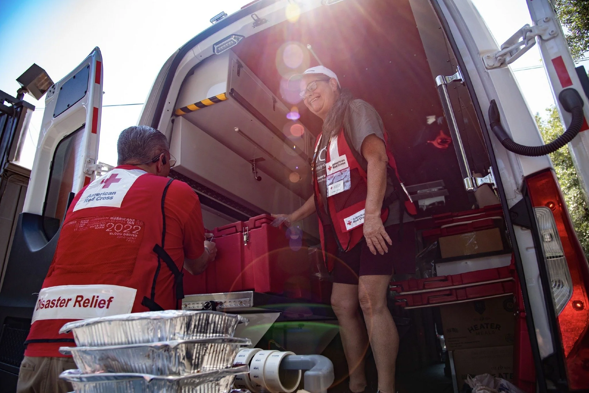 Two American Red Cross workers inside a mobile blood donation unit, one woman smiling and handing a blood donation kit to a man, with sun glare and trees outside.