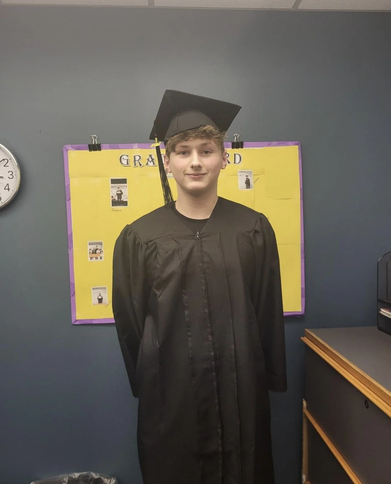 A young man in a black graduation gown and cap standing in front of a yellow bulletin board with purple border, in a room with a wall clock on the left side.