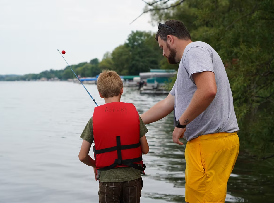 A man helps a boy fish at a lake