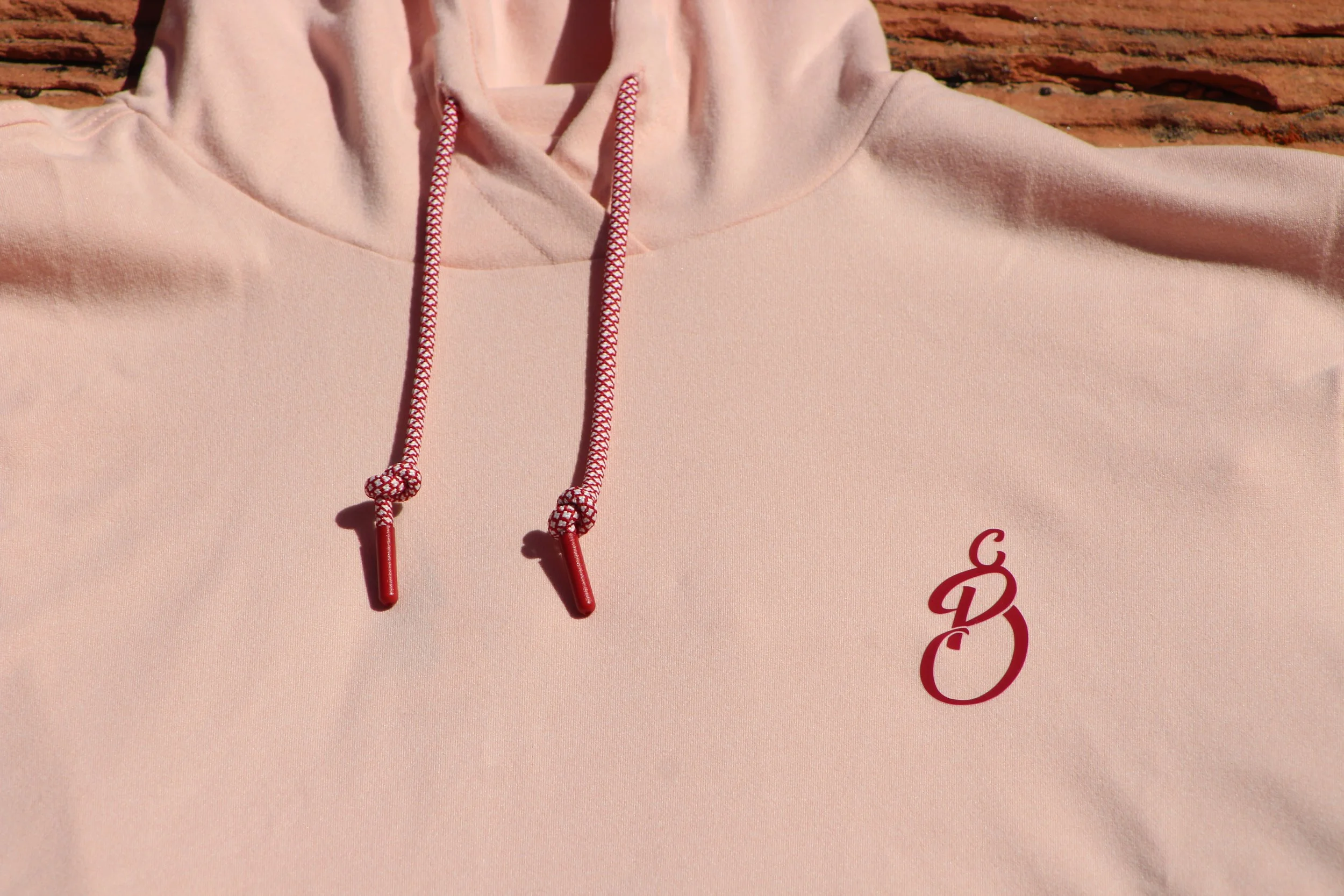 Close-up of a light pink hoodie with red and pink drawstrings and a small red logo on the chest area.