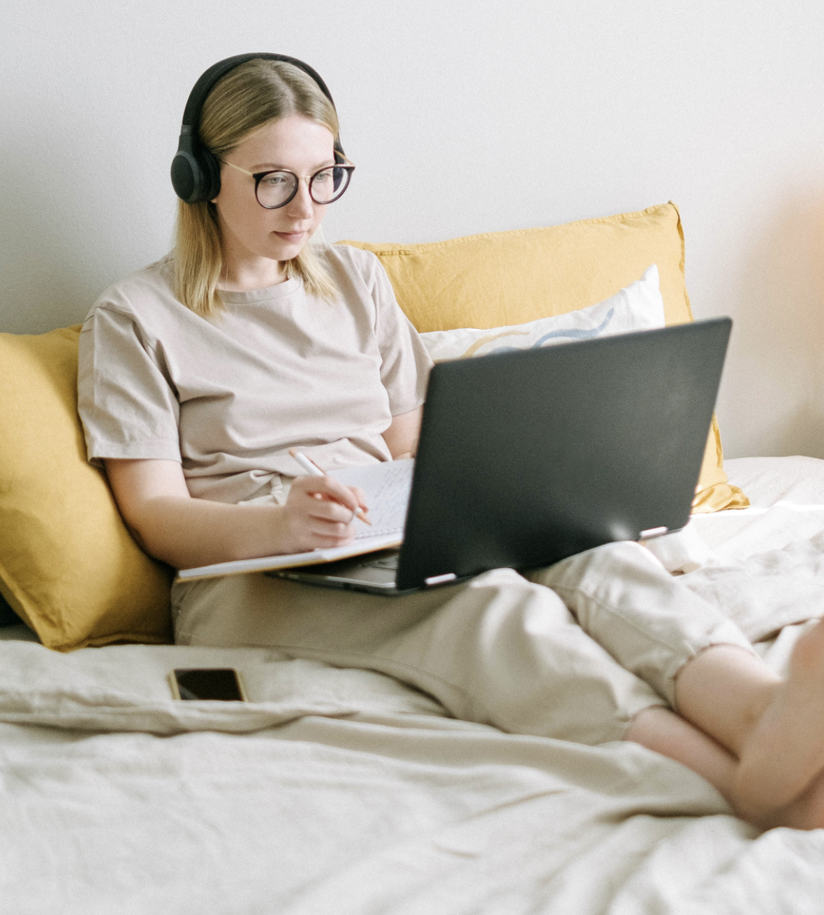Young woman with glasses and headphones sitting on bed with yellow pillows, using a laptop and taking notes with a pen, smartphone on bed nearby.