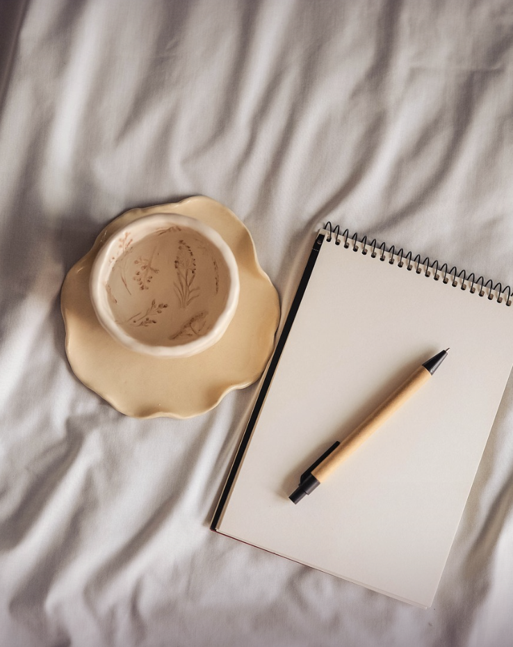 A sketchbook with a pen resting on it, and a ring dish on a soft fabric surface.