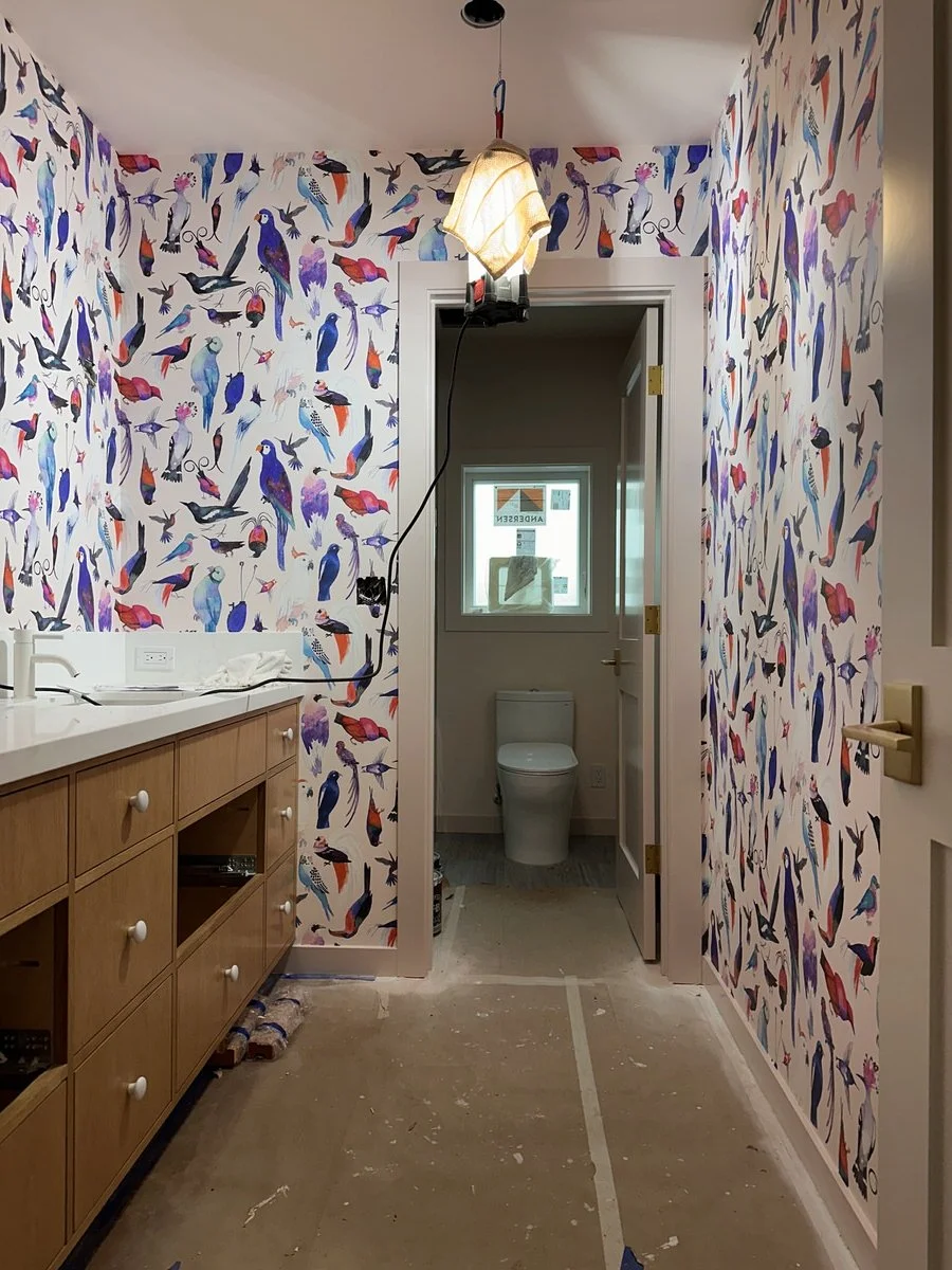 Kids' bathroom during wallpaper installation showing colorful bird-print wallpaper in progress, oak vanity, and separate water closet