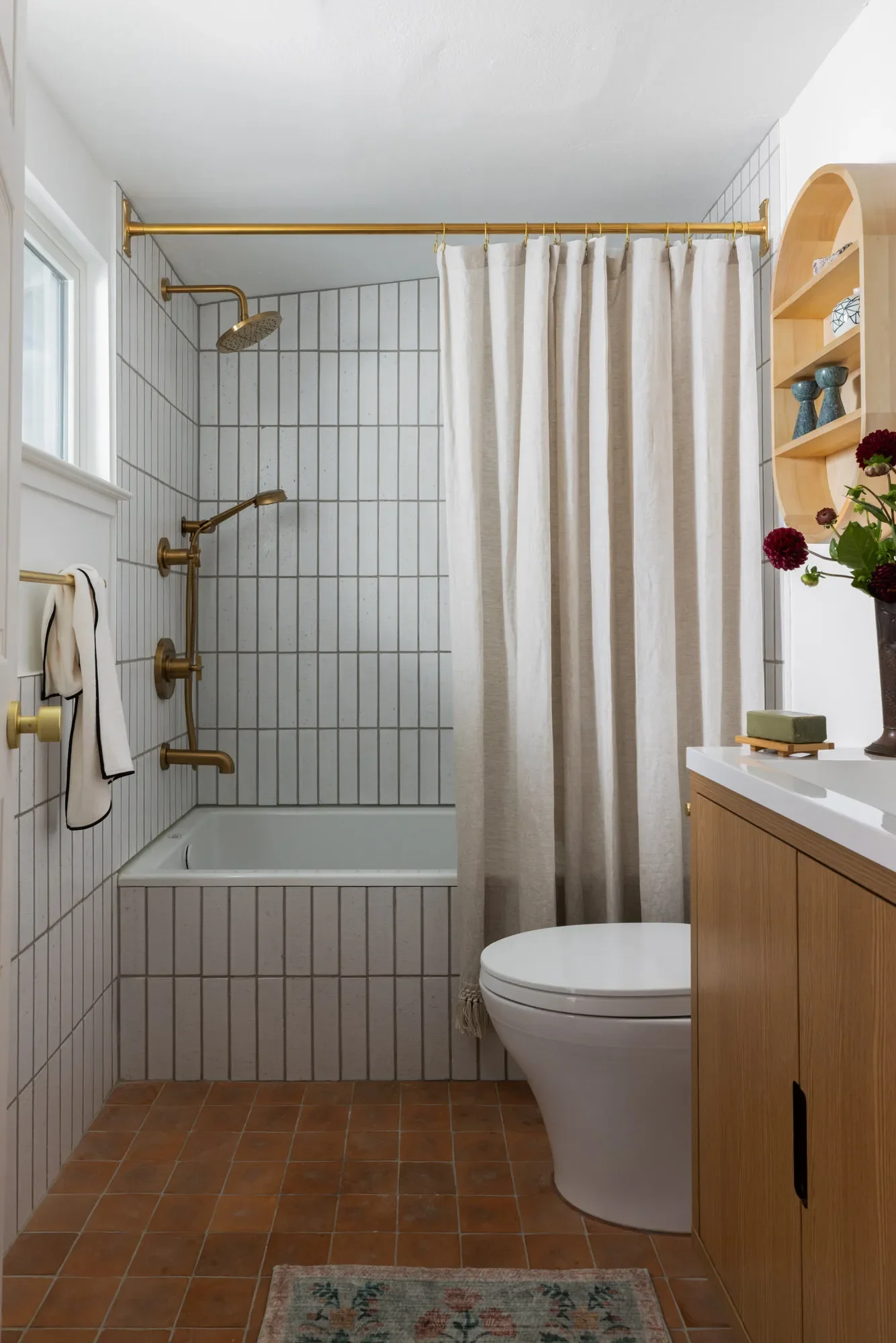 Compact bathroom with white vertical tile, brass shower fixtures, a linen curtain, terracotta floors, an oak vanity, and dahlias in a vase.