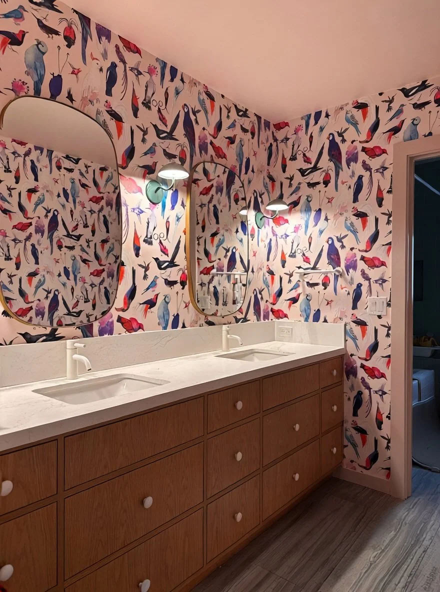Completed kids' bathroom with floor-to-ceiling colorful bird wallpaper, double undermount sinks on a warm oak vanity, white stone countertop, and brass oval mirrors with wall sconces