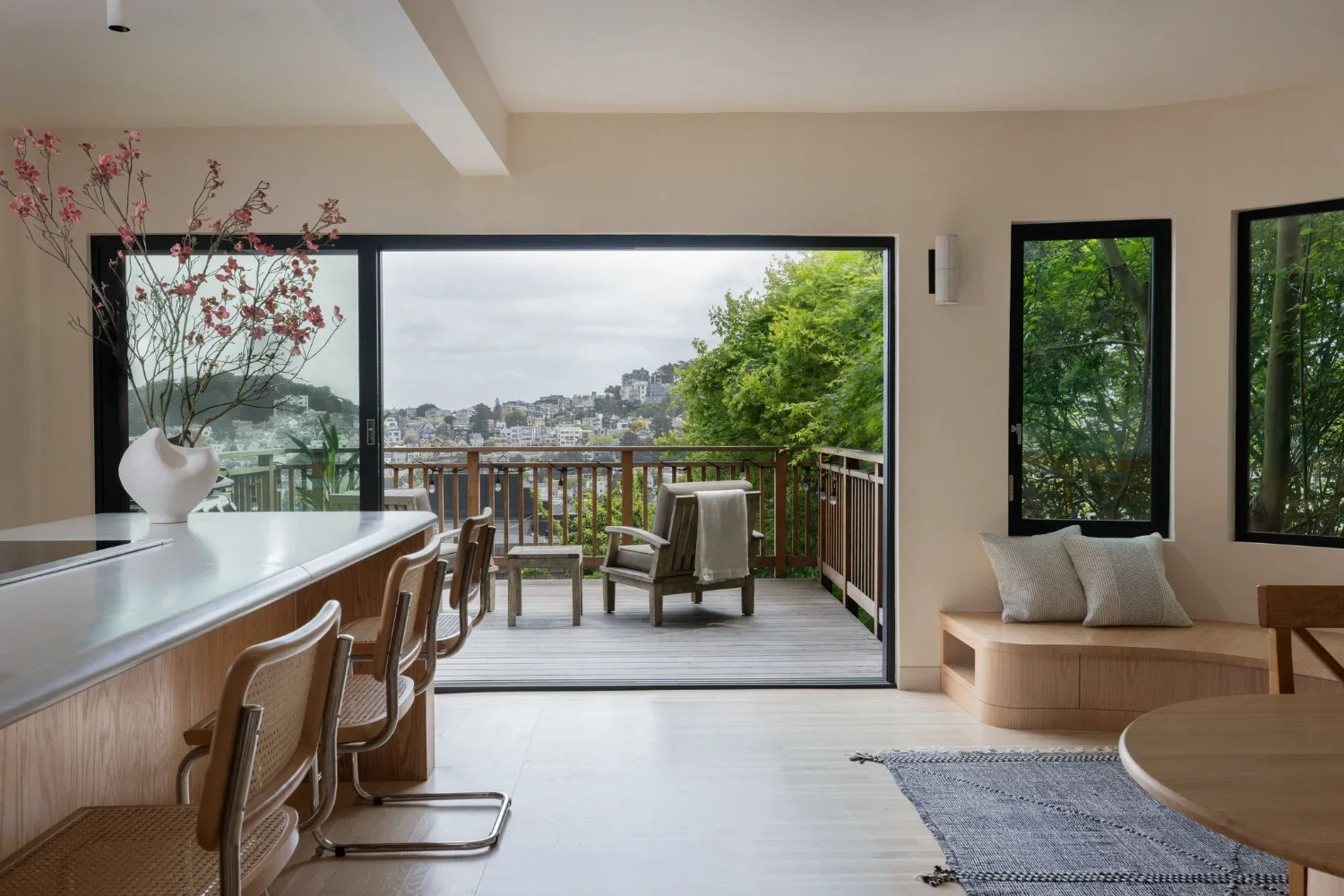 Warm minimalist San Francisco home interior featuring red oak kitchen island, cane cantilever bar stools, sliding black-frame glass doors opening to a deck with city views — Studio VAE interior design