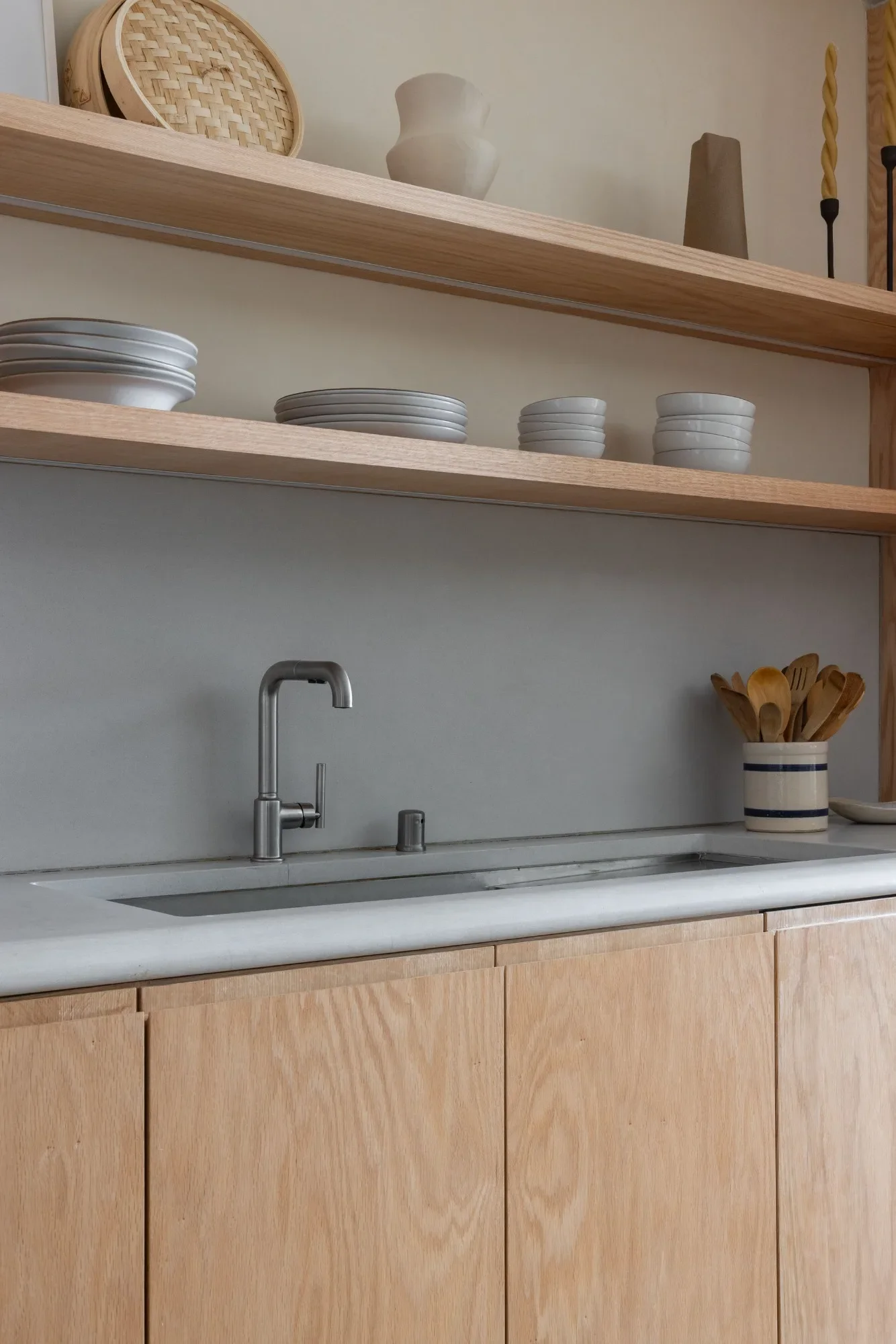 Open oak shelves holding stacked white dishware, with a woven basket, ceramic vases, and twisted taper candles displayed above.